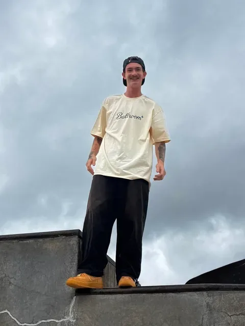 Trenton standing on top of a concrete ledge in the Ballroom tee, smiling