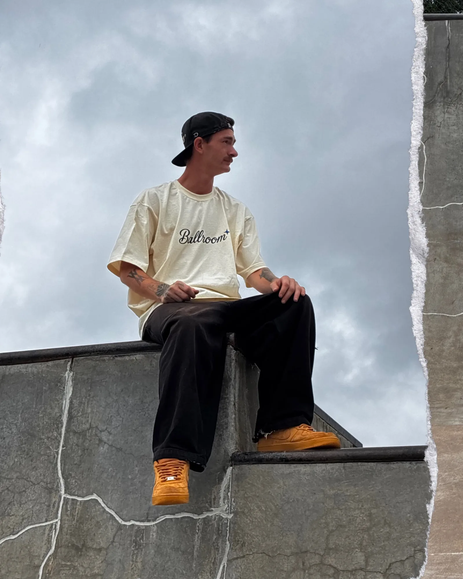 Trenton sitting on a skatepark ledge in the Ballroom tee under a cloudy sky