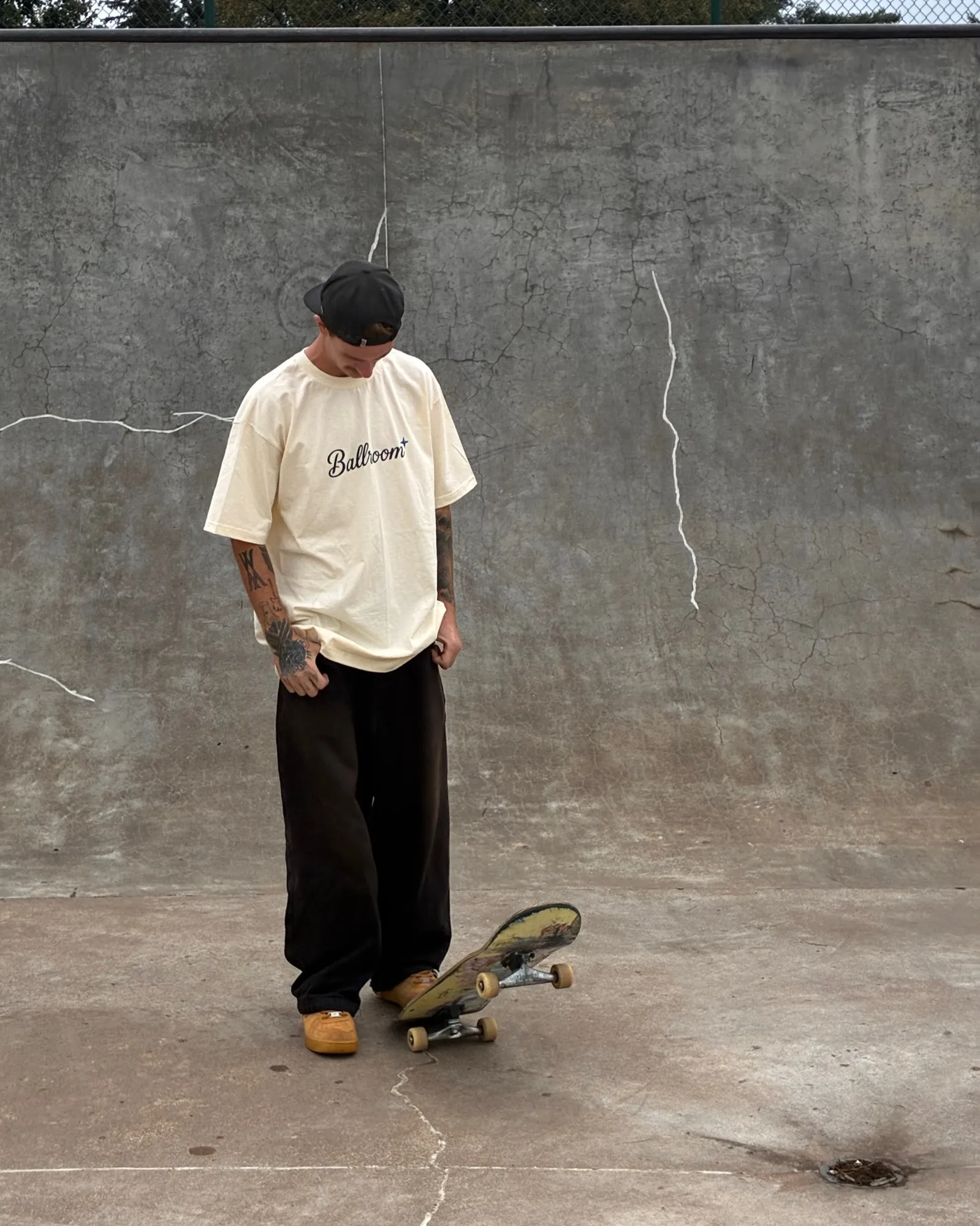 Trenton standing in a concrete bowl in the Ballroom tee, looking down at his board